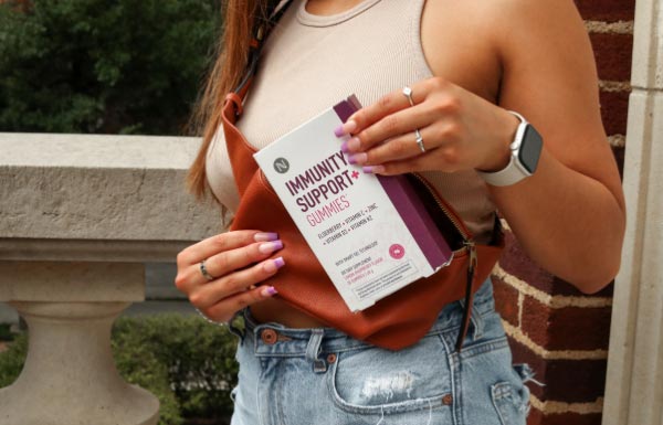 A woman outside in a summery outfit holding a box of Immunity Support+ Gummies to her chest.